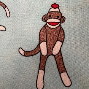 May include: A repeating pattern of sock monkey illustrations on a light blue background. Each monkey is brown with white accents on the hands and feet, a red mouth, and a red flower on its hat.