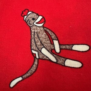 May include: A sock monkey design on a red background. The sock monkey is brown with white accents on the hands, feet, and face. It has a red mouth and a red hat. The background is a solid, bright red.