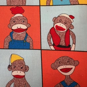 May include: Fabric featuring a repeating pattern of sock monkey illustrations. Each monkey has a brown body, white face, and red mouth. The background is divided into colorful squares of orange, yellow, and light blue.