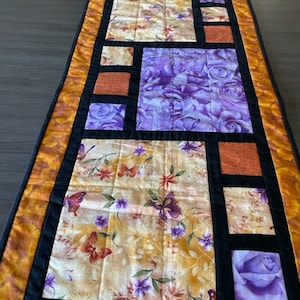 May include: A quilted table runner with a patchwork design featuring purple roses, butterflies, and orange and yellow floral patterns. The runner has a black border and a gold trim.