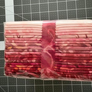 May include: A stack of fabric strips in a gradient of pink and red shades, wrapped in clear plastic. The fabric features various tonal patterns and is positioned over a black cutting mat with white grid lines showing measurements in inches.
