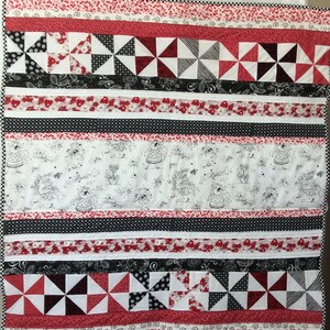 May include: A red, white, and black quilt with a patchwork design. The quilt features a variety of fabrics, including floral prints, polka dots, and geometric patterns. The quilt is made up of several rows of fabric, each with a different design. The quilt is edged with a white trim.