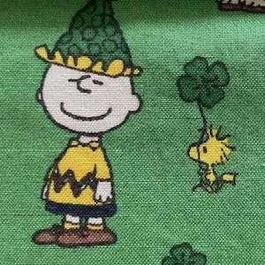 May include: A green fabric with a cartoon image of Charlie Brown wearing a green shamrock hat and a yellow shirt with a black zig-zag pattern.  A yellow bird with a green shamrock is flying next to Charlie Brown.  There are several green shamrocks scattered throughout the fabric.