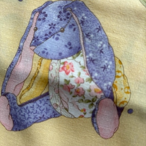 May include: A fabric bunny with a patchwork design. The bunny has a lavender head and ears with a floral pattern, pink accents, and a white floral belly. The background is a pale yellow.