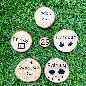 May include: Six wooden circles with black text on a green grass background. The circles say "Today is...", "Friday", "29", "October", "The Weather is...", and "Raining".