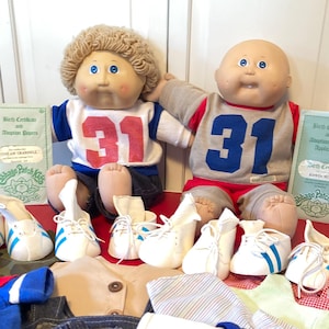 May include: Two Cabbage Patch Kids dolls wearing football jerseys with the number 31. The dolls are surrounded by various outfits, including denim overalls, shirts, and white sneakers with blue stripes. Birth certificates are also present.
