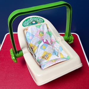 Könnte beinhalten: Ein Vintage-Babysitz von Cabbage Patch Kids in Creme und Grün. Der Sitz hat einen grünen Griff und einen Innenraum aus gemustertem Stoff mit Cabbage Patch Kids-Figuren. Der Sitz steht auf einer roten Oberfläche.