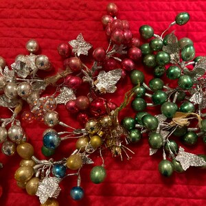 Puede incluir: Primer plano de adornos navideños vintage en varios colores, incluyendo plata, rojo, verde, dorado y azul. Los adornos tienen forma de bayas y están unidos a tallos de alambre plateado con hojas metálicas. El fondo es de tela roja.
