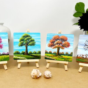 May include: Four small paintings on easels depicting a tree in each of the four seasons. The paintings are on a brown surface with two seashells in front of them.