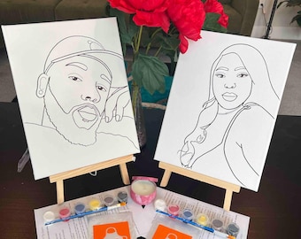 May include: Two white canvases on wooden easels featuring black line art portraits of a person wearing a baseball cap and a person with long hair. Includes two sets of paint pots, two paintbrushes, two instruction sheets, and two aprons.