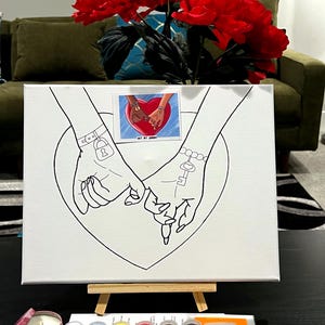 May include: A white canvas with a black line drawing of two hands holding each other. The hands are intertwined and have a key and a lock on their wrists. The canvas is on an easel and there are paint pots and a paintbrush in the foreground. The text "Art By Urban" is visible on the canvas.
