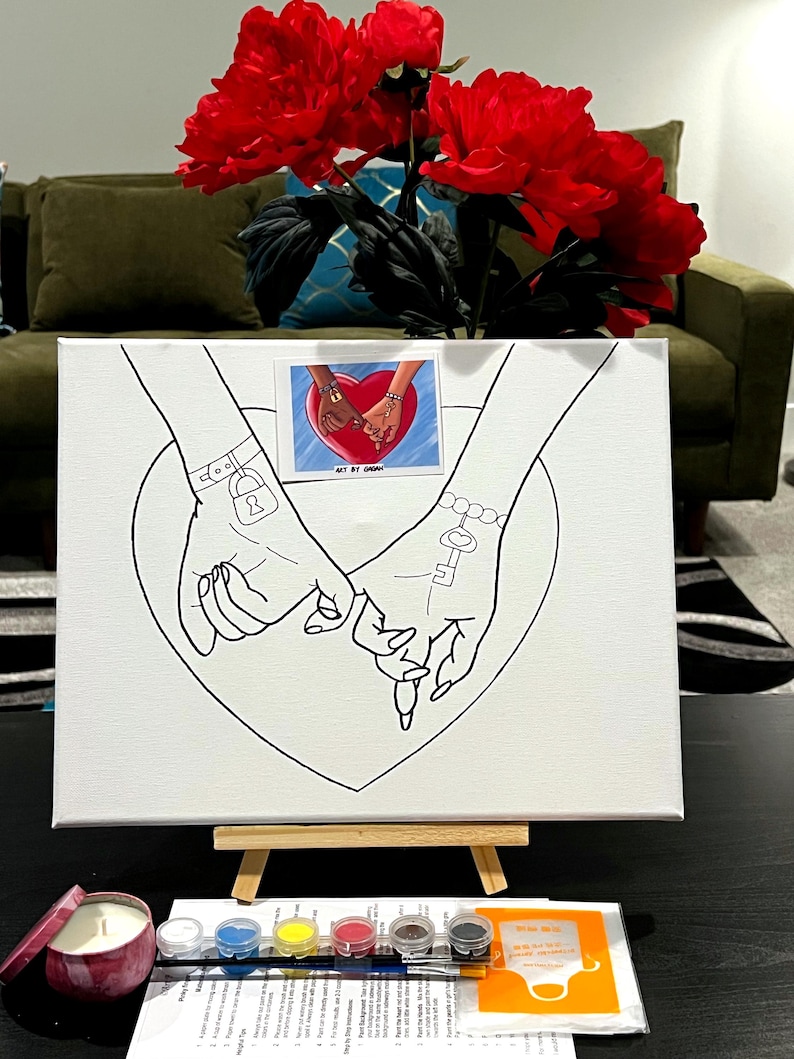 May include: A white canvas with a black line drawing of two hands holding each other. The hands are intertwined and have a key and a lock on their wrists. The canvas is on an easel and there are paint pots and a paintbrush in the foreground. The text "Art By Urban" is visible on the canvas.
