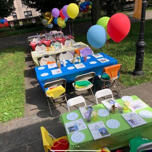 May include: A party setup with multiple tables covered in colorful tablecloths. Each table has white plates, paintbrushes, and canvases. There are colorful balloons in the background.
