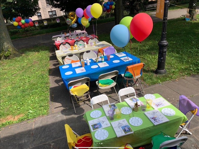 May include: A party setup with multiple tables covered in colorful tablecloths. Each table has white plates, paintbrushes, and canvases. There are colorful balloons in the background.