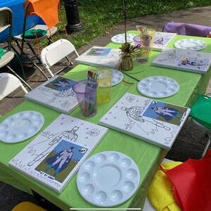 May include: A green tablecloth with four paint-by-number canvases featuring a superhero character. Each canvas has a white paint palette with 12 wells. There are also two purple cups with paintbrushes and a gold decorative piece on the table.