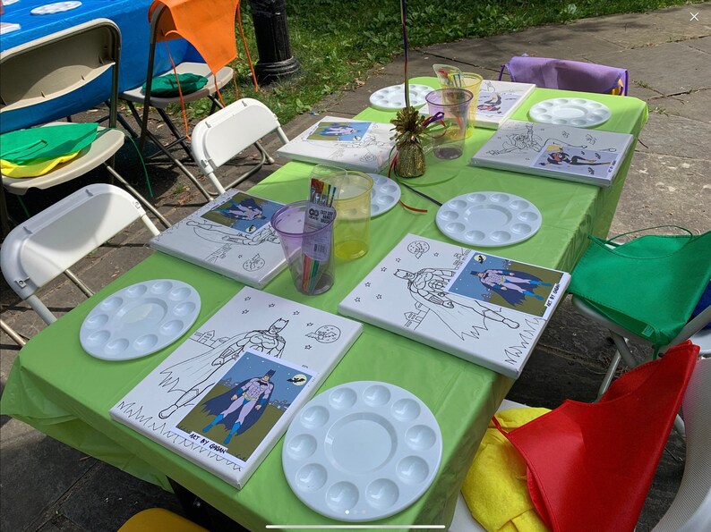 May include: A green tablecloth with four paint-by-number canvases featuring a superhero character. Each canvas has a white paint palette with 12 wells. There are also two purple cups with paintbrushes and a gold decorative piece on the table.