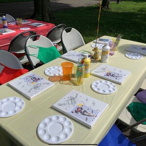 May include: A table set up for a painting party with white canvases, paint palettes, paint bottles, and paintbrushes. There are also red, green, and blue aprons on the chairs.