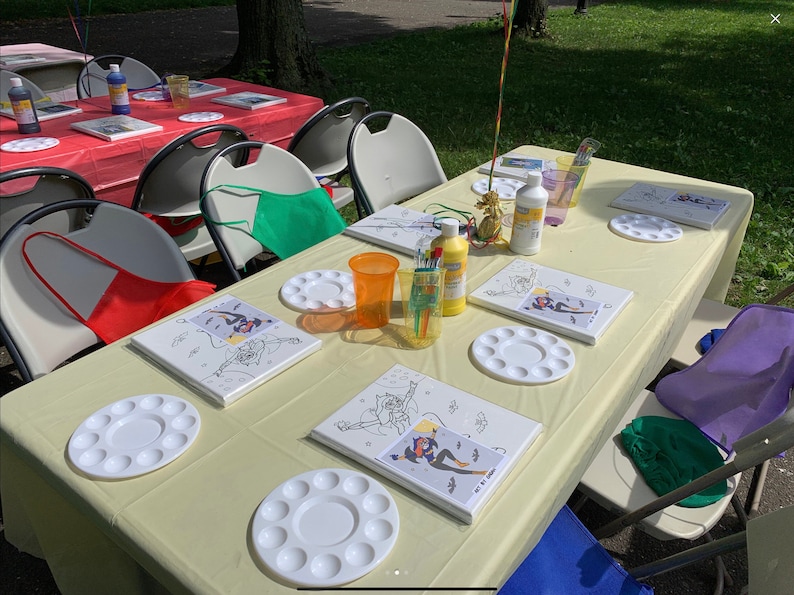 May include: A table set up for a painting party with white canvases, paint palettes, paint bottles, and paintbrushes. There are also red, green, and blue aprons on the chairs.