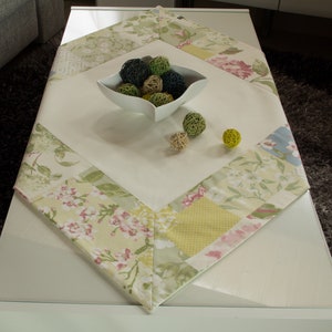 Op de afbeelding: Een wit tafelkleed met een bloemig patchwork-ontwerp in tinten groen, roze en wit. Het tafelkleed ligt op een witte tafel met een kom met decoratieve ballen in het midden.