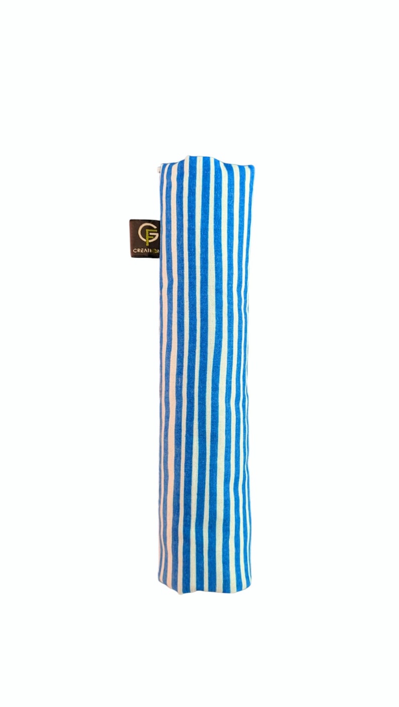 Puede incluir: Un estuche cil&iacute;ndrico de tela con un patr&oacute;n de rayas verticales azules y blancas. Una peque&ntilde;a etiqueta rectangular de color marr&oacute;n oscuro est&aacute; unida al lateral, mostrando un logotipo dorado con la letra G y el texto CREATIONS impreso debajo.