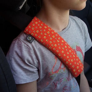 Puede incluir: Una funda rectangular para cintur&oacute;n de seguridad hecha de tela naranja vibrante con un patr&oacute;n repetitivo de peque&ntilde;os c&iacute;rculos amarillos con centros oscuros. La funda est&aacute; asegurada sobre un cintur&oacute;n de seguridad negro y tiene una peque&ntilde;a etiqueta negra con un logotipo blanco.