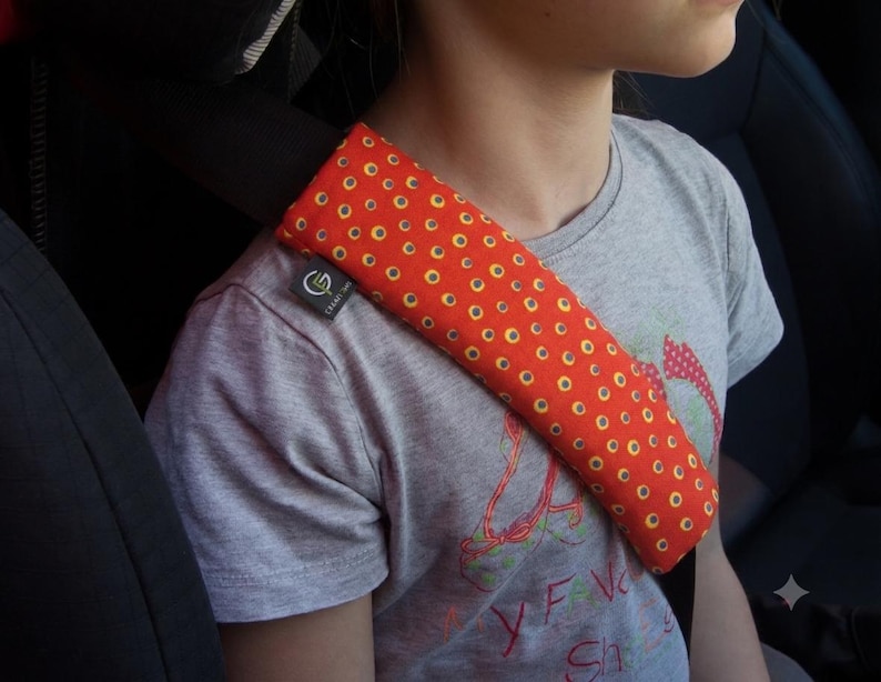 Puede incluir: Una funda rectangular para cintur&oacute;n de seguridad hecha de tela naranja vibrante con un patr&oacute;n repetitivo de peque&ntilde;os c&iacute;rculos amarillos con centros oscuros. La funda est&aacute; asegurada sobre un cintur&oacute;n de seguridad negro y tiene una peque&ntilde;a etiqueta negra con un logotipo blanco.