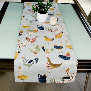 May include: A grey table runner with a repeating pattern of colourful chickens. The runner is on a glass table with a white candle and a potted plant.