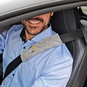 Könnte beinhalten: Ein Mann trägt ein hellblaues Hemd mit einem grauen Sicherheitsgurtpolster, auf dem "Smart" in weißen Buchstaben steht. Der Sicherheitsgurt ist schwarz und das Polster hat ein gelbes Dreieck mit einem stilisierten "C" darin.