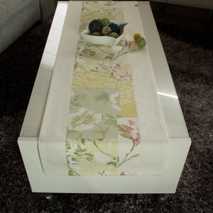 Può includere: Un runner da tavolo bianco con un motivo floreale in tonalità di verde, giallo e rosa. Il runner è su un tavolino da caffè bianco con una ciotola di palline decorative verdi e marroni.