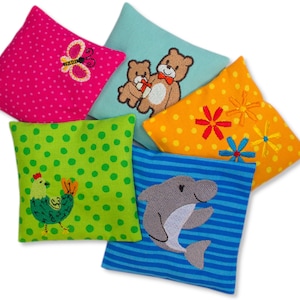 Könnte beinhalten: Fünf bunte quadratische Kissen mit gestickten Designs. Die Kissen zeigen einen Schmetterling, zwei Bären, Blumen, ein Huhn und einen Delfin.