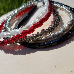 Könnte beinhalten: Ein Set aus fünf Perlenarmbändern in Schwarz, Weiß, Grau und Rot. Die Armbänder sind übereinander gestapelt.