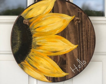 May include: A round wooden door hanger with a painted sunflower design. The sunflower is yellow with a dark brown center. The words "Hello" are written in white on the wood.