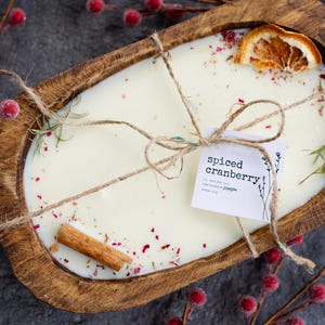 May include: A white candle in a wooden bowl with a cinnamon stick, dried orange slices, and red sprinkles. The candle is tied with twine and a tag that reads "Spiced Cranberry".