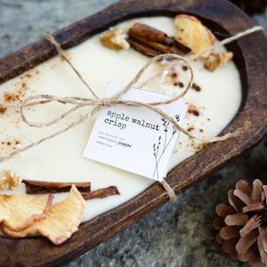 May include: A wooden bowl candle with a white wax base and a label that reads "apple walnut crisp". The candle is decorated with dried apple slices, cinnamon sticks, and a brown twine bow.