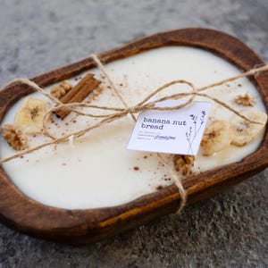 May include: A banana nut bread scented candle in a rustic wooden bowl. The candle is white and decorated with banana slices, walnuts, and a cinnamon stick. A twine bow and a tag that reads "banana nut bread" complete the design.