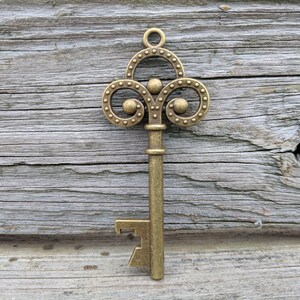 May include: A gold-toned metal key with a decorative, ornate top and a bottle opener on the bottom. The key is against a weathered wooden background.