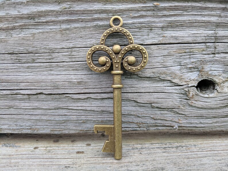 May include: A gold-toned metal key with a decorative, ornate top and a bottle opener on the bottom. The key is against a weathered wooden background.