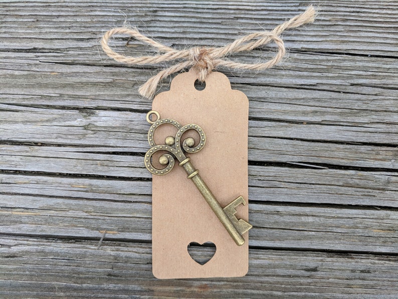 May include: A brown paper tag with a heart-shaped cutout and a decorative key-shaped bottle opener attached. The tag is tied with twine.