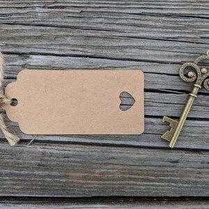 May include: A brown paper gift tag with a heart-shaped cutout tied with twine on a rustic wooden background. A gold-toned decorative key is also on the wood.