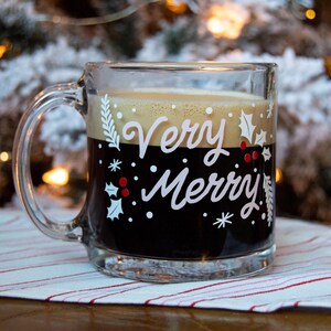 Puede incluir: Una taza de vidrio transparente con una bebida de caf&eacute; negro en su interior. La taza tiene un dise&ntilde;o blanco con las palabras "Very Merry" y hojas de acebo.