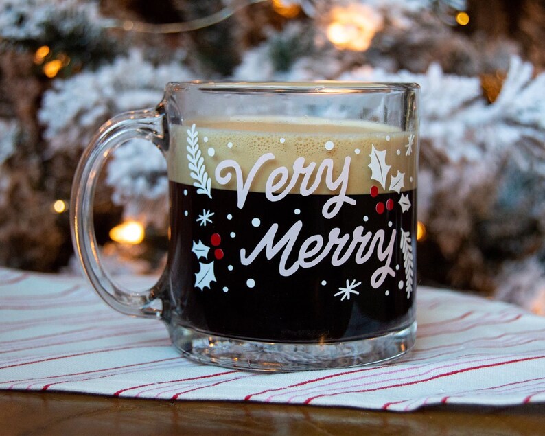Puede incluir: Una taza de vidrio transparente con una bebida de caf&eacute; negro en su interior. La taza tiene un dise&ntilde;o blanco con las palabras "Very Merry" y hojas de acebo.