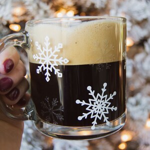 May include: A clear glass mug filled with dark coffee and topped with foam. The mug features white snowflake designs. The mug is held by a person with dark red nail polish.