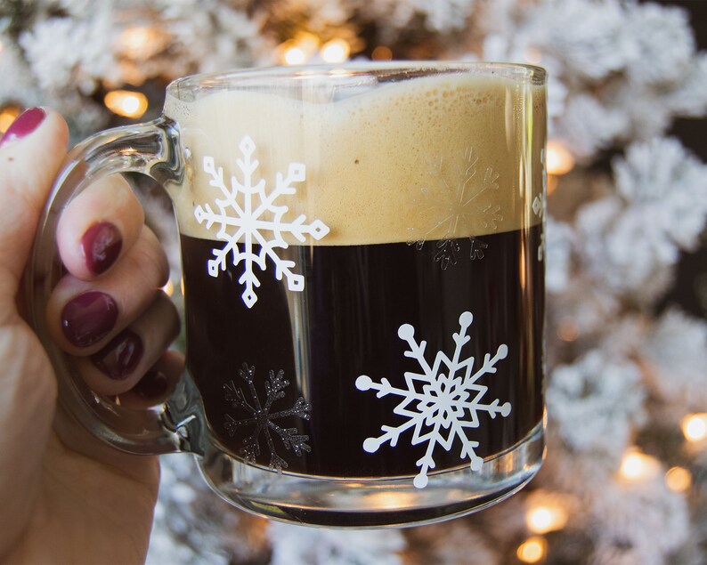May include: A clear glass mug filled with dark coffee and topped with foam. The mug features white snowflake designs. The mug is held by a person with dark red nail polish.