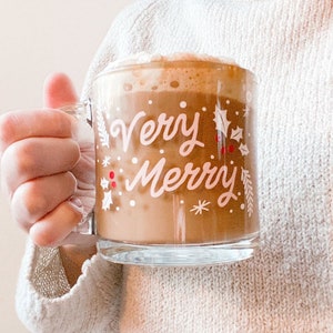 Puede incluir: Una taza de vidrio con fondo blanco y las palabras "Very Merry" en letras rosas. La taza est&aacute; llena de una bebida marr&oacute;n cubierta con crema batida.