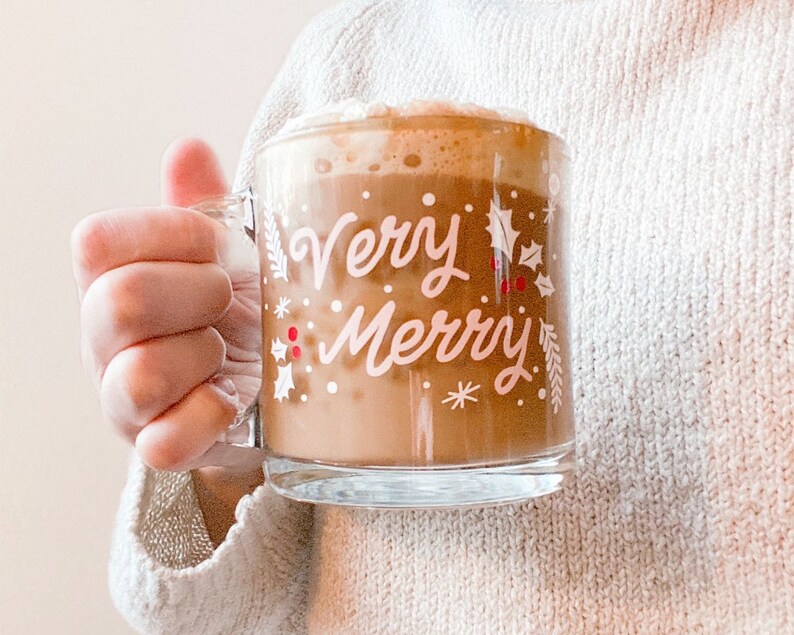 Puede incluir: Una taza de vidrio con fondo blanco y las palabras "Very Merry" en letras rosas. La taza est&aacute; llena de una bebida marr&oacute;n cubierta con crema batida.