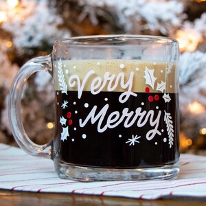 Puede incluir: Una taza de vidrio transparente con asa negra llena de caf&eacute;. La taza tiene un dise&ntilde;o blanco con las palabras "Very Merry" y una rama de acebo con bayas rojas.