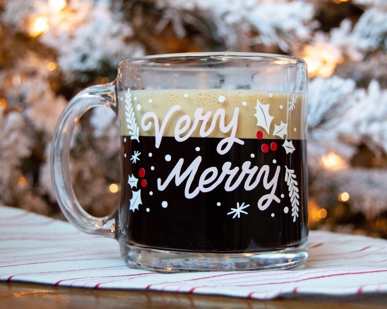 Puede incluir: Una taza de vidrio transparente con asa negra llena de caf&eacute;. La taza tiene un dise&ntilde;o blanco con las palabras "Very Merry" y una rama de acebo con bayas rojas.