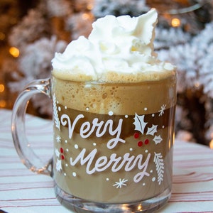 Puede incluir: Una taza de vidrio con asa blanca llena de una bebida de caf&eacute; coronada con crema batida. La taza tiene un dise&ntilde;o blanco que dice "Very Merry" con acebo y bayas.
