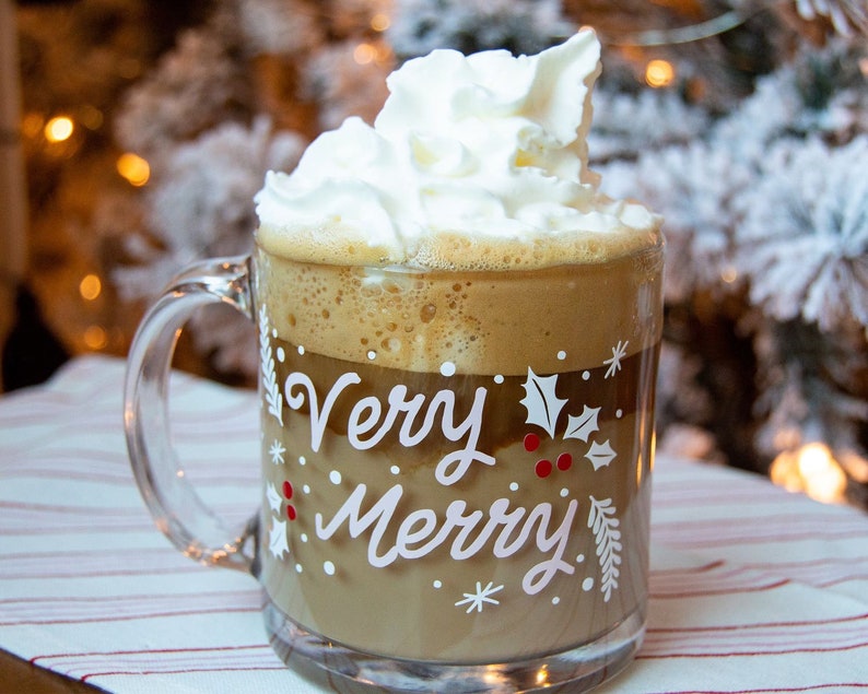 Puede incluir: Una taza de vidrio con asa blanca llena de una bebida de caf&eacute; coronada con crema batida. La taza tiene un dise&ntilde;o blanco que dice "Very Merry" con acebo y bayas.