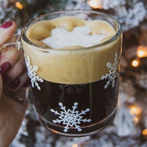 May include: A clear glass mug filled with dark coffee and topped with foam and sugar. The mug features white snowflake designs. A hand with red nail polish holds the mug. The background is blurred with Christmas lights.